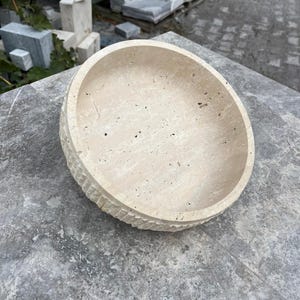 K&ouml;nnte beinhalten: Eine beige Steinschale mit strukturierter Au&szlig;enseite und glatter Innenseite. Die Schale ist rund, flach und steht auf einer grauen Oberfl&auml;che. Die Au&szlig;enseite hat ein geschnitztes, geriffeltes Muster.