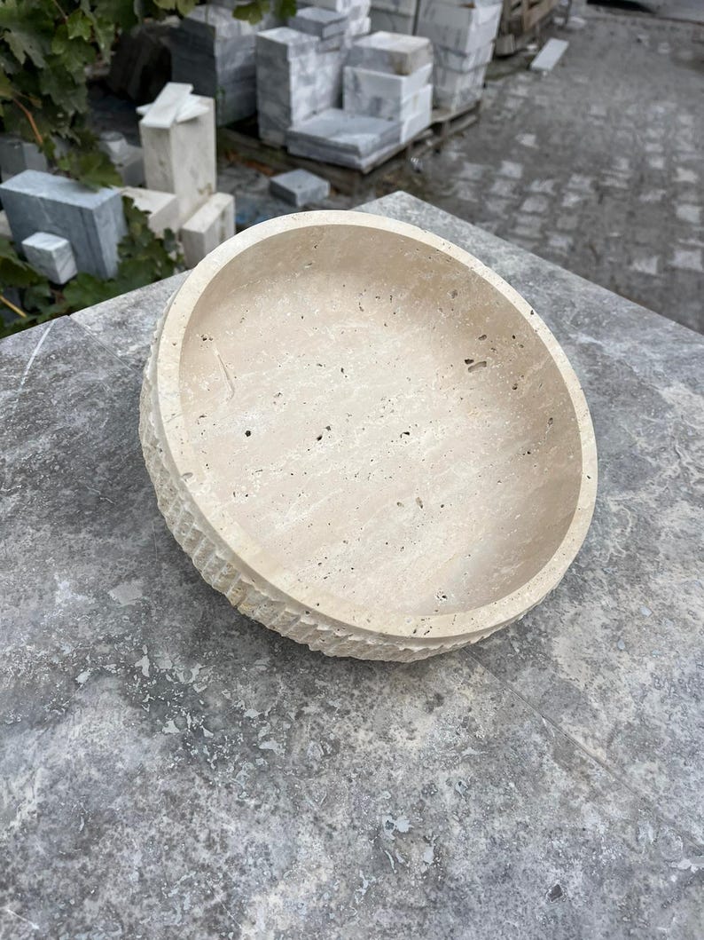 K&ouml;nnte beinhalten: Eine beige Steinschale mit strukturierter Au&szlig;enseite und glatter Innenseite. Die Schale ist rund, flach und steht auf einer grauen Oberfl&auml;che. Die Au&szlig;enseite hat ein geschnitztes, geriffeltes Muster.