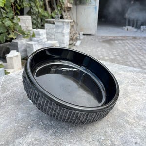 May include: A black, round bowl with a textured, ridged exterior. The bowl is made of a glossy material and sits on a grey, stone surface. The bowl's interior is smooth and reflective.