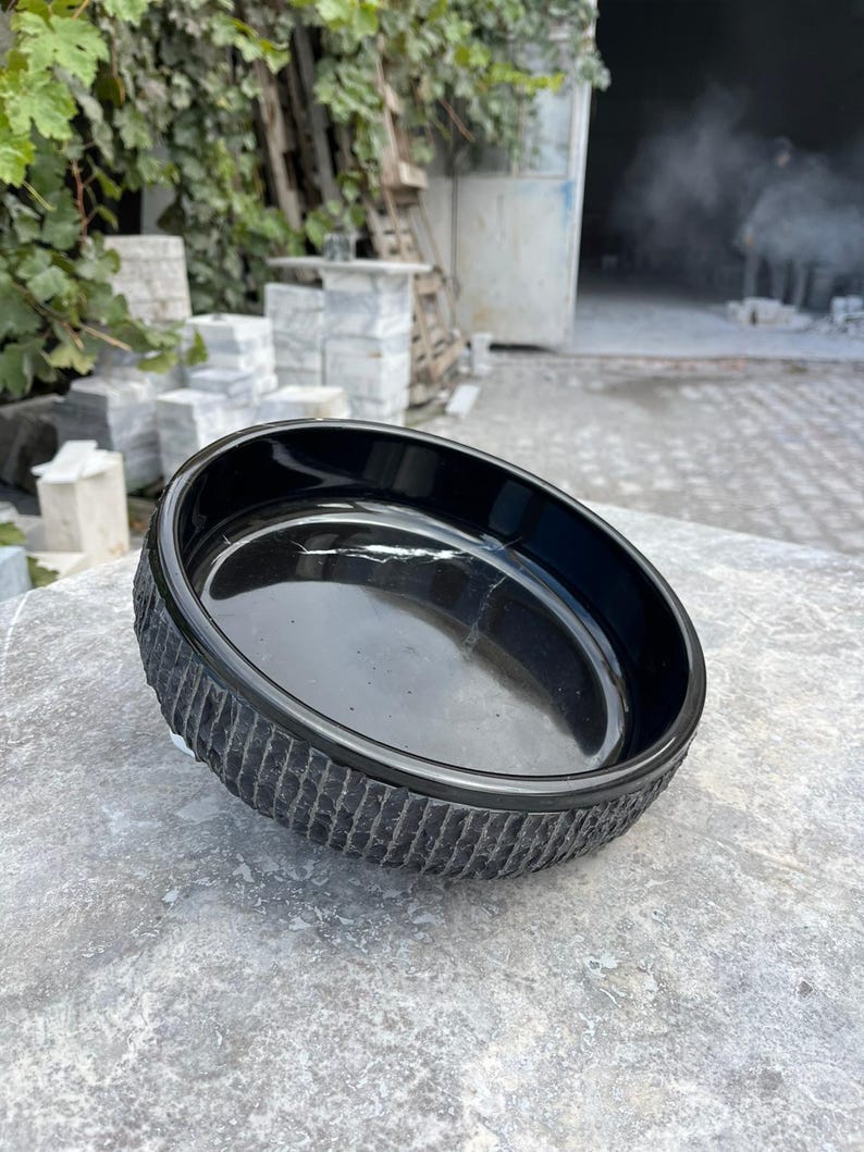 May include: A black, round bowl with a textured, ridged exterior. The bowl is made of a glossy material and sits on a grey, stone surface. The bowl's interior is smooth and reflective.