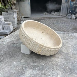 K&ouml;nnte beinhalten: Eine beige, ovale Steinschale mit einer strukturierten, geriffelten Au&szlig;enseite. Die Schale ruht auf einem kleinen, quadratischen, hellfarbigen Steinblock. Der Hintergrund zeigt eine Werkstatteinstellung mit Steinbl&ouml;cken und einer offenen T&uuml;r.