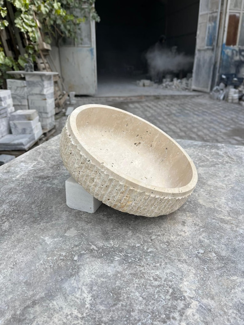 K&ouml;nnte beinhalten: Eine beige, ovale Steinschale mit einer strukturierten, geriffelten Au&szlig;enseite. Die Schale ruht auf einem kleinen, quadratischen, hellfarbigen Steinblock. Der Hintergrund zeigt eine Werkstatteinstellung mit Steinbl&ouml;cken und einer offenen T&uuml;r.