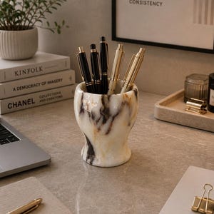 May include: A cylindrical pen holder crafted from white marble with prominent black and brown veining. The holder contains three black pens with gold-toned clips and two metallic gold pens, all standing upright within the vessel's smooth interior.