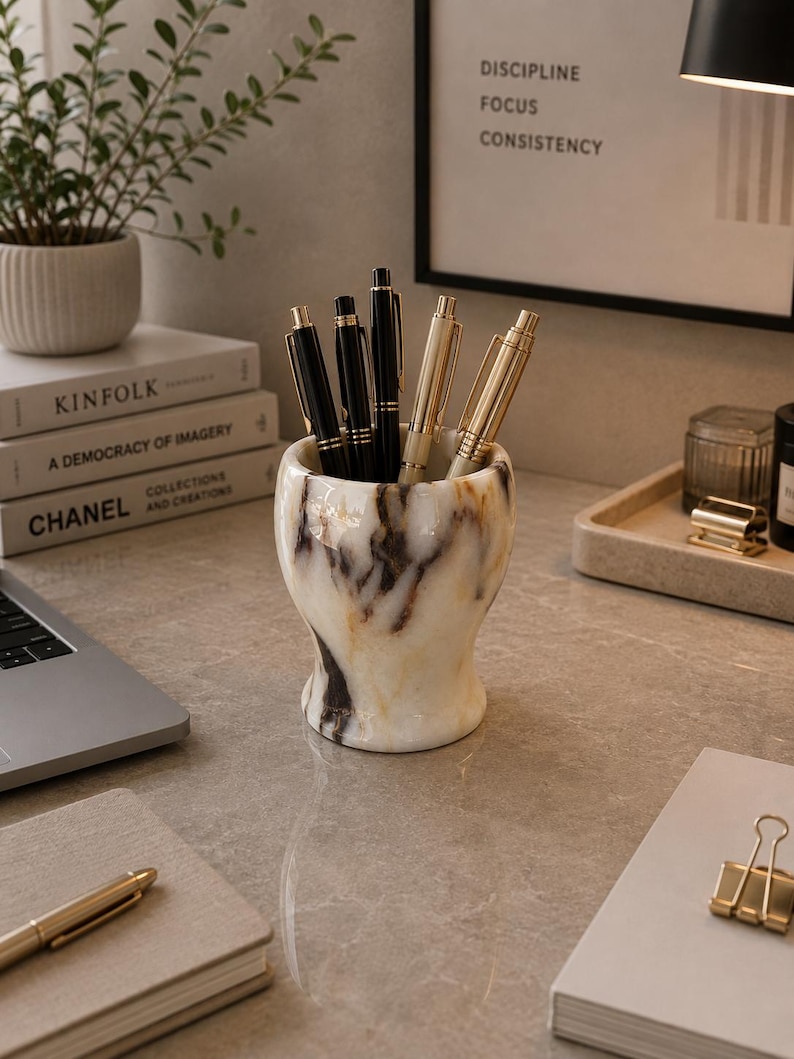 May include: A cylindrical pen holder crafted from white marble with prominent black and brown veining. The holder contains three black pens with gold-toned clips and two metallic gold pens, all standing upright within the vessel's smooth interior.