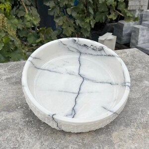 May include: A round, white marble bowl with black veining. The bowl has a textured rim and a smooth interior. The natural stone bowl is ideal for decorative use or as a serving dish. The bowl is approximately 25 cm in diameter.