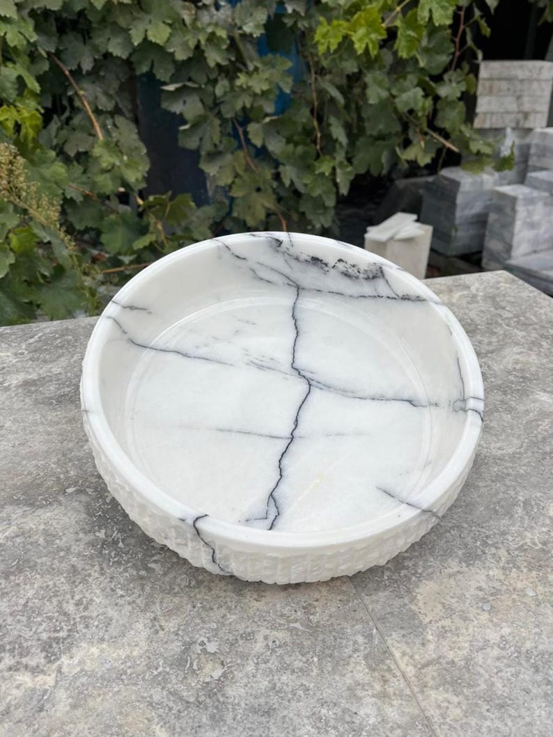 May include: A round, white marble bowl with black veining. The bowl has a textured rim and a smooth interior. The natural stone bowl is ideal for decorative use or as a serving dish. The bowl is approximately 25 cm in diameter.