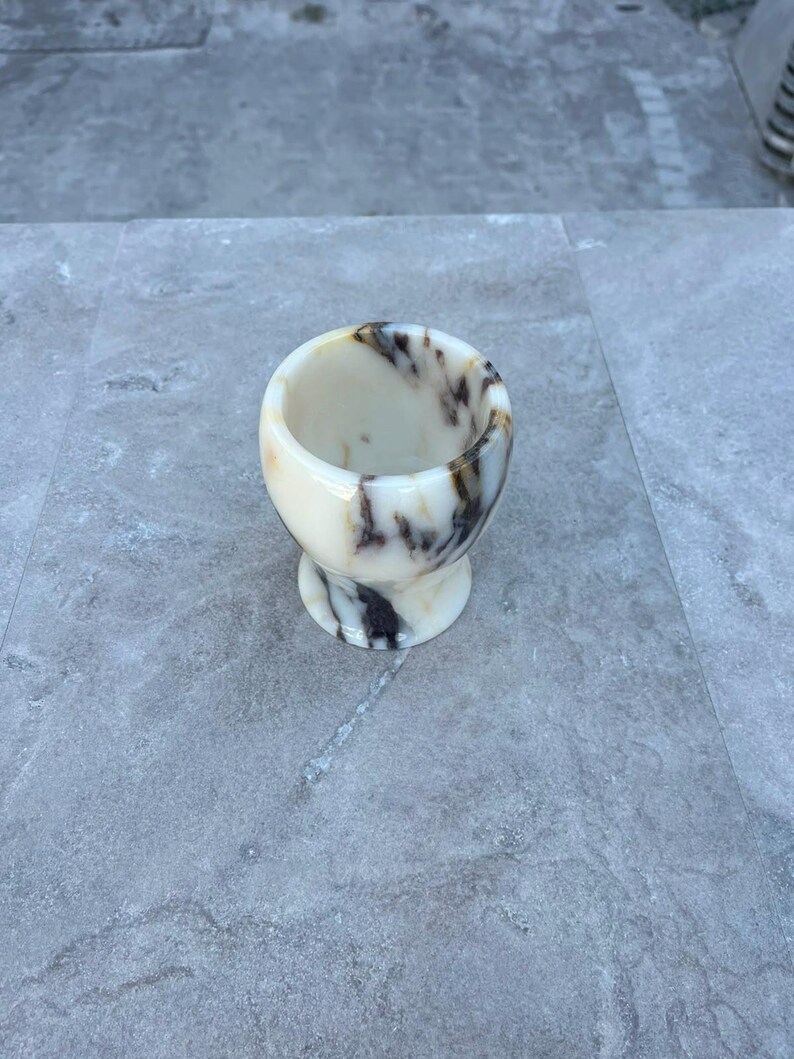 May include: A small, rounded cup crafted from polished stone, featuring a smooth, cream-coloured base with prominent, swirling dark brown and black veins throughout the material. The vessel has a wide, open top and a narrow, stable circular pedestal base.
