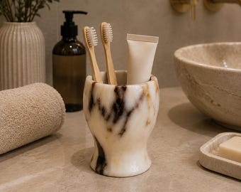 May include: A cylindrical marble toothbrush holder with a flared base and rim, featuring a polished white surface with prominent dark brown and tan veining. The holder contains two light-colored wooden toothbrushes and a plain beige tube of toothpaste.