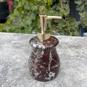 Puede incluir: Un dispensador de jab&oacute;n decorativo con una bomba dorada. El dispensador est&aacute; hecho de m&aacute;rmol rojo oscuro con vetas blancas. El dispensador tiene una forma redondeada y se encuentra sobre una superficie gris.