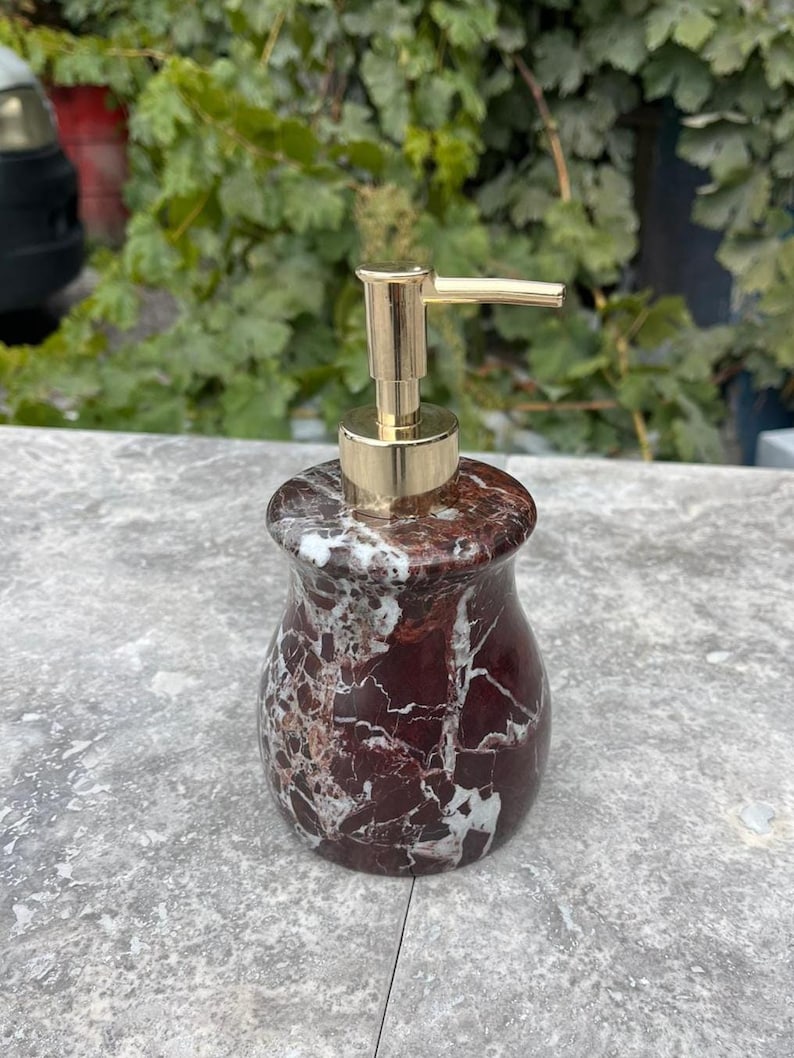 Puede incluir: Un dispensador de jab&oacute;n decorativo con una bomba dorada. El dispensador est&aacute; hecho de m&aacute;rmol rojo oscuro con vetas blancas. El dispensador tiene una forma redondeada y se encuentra sobre una superficie gris.