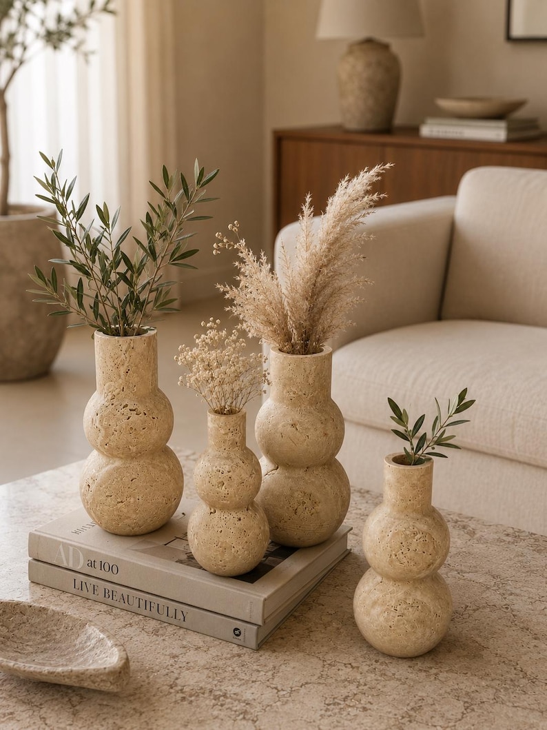Puede incluir: Cuatro jarrones de travertino beige con una silueta apilada de doble esfera. Los jarrones presentan una textura de piedra natural porosa y contienen varios tallos bot&aacute;nicos secos. Est&aacute;n dispuestos sobre un libro titulado AD at 100 LIVE BEAUTIFULLY.