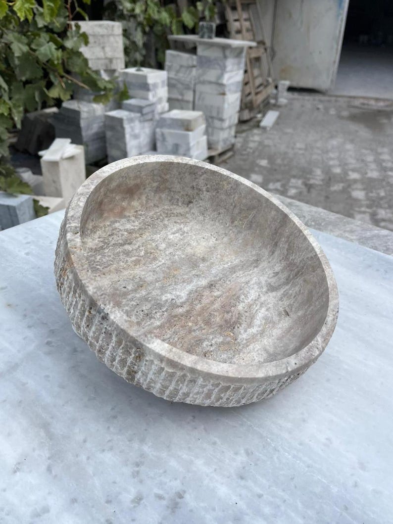 May include: A round, stone bowl with a textured exterior and a smooth interior. The bowl is a light beige color with darker brown and gray veining. The bowl is sitting on a white marble surface.