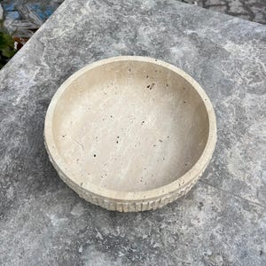 K&ouml;nnte beinhalten: Eine beige Steinschale mit runder Form und strukturierter, geriffelter Au&szlig;enseite. Die Schale steht auf einer grauen Steinoberfl&auml;che. Das Innere der Schale ist glatt und hell.