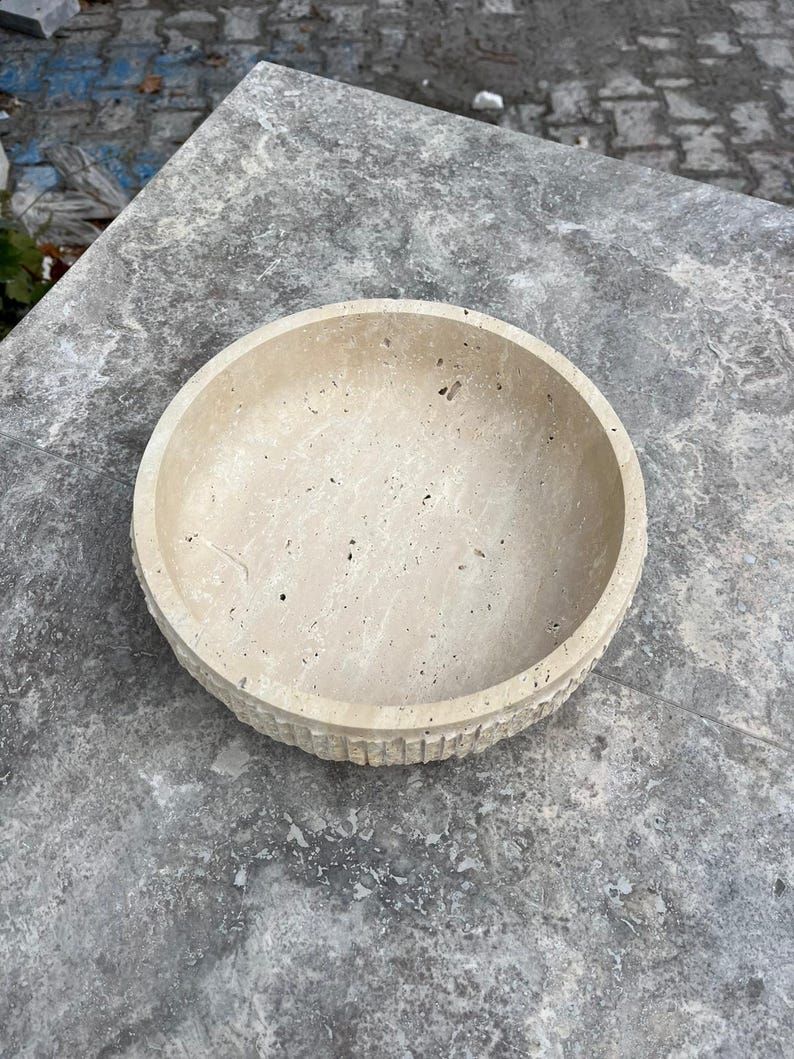 K&ouml;nnte beinhalten: Eine beige Steinschale mit runder Form und strukturierter, geriffelter Au&szlig;enseite. Die Schale steht auf einer grauen Steinoberfl&auml;che. Das Innere der Schale ist glatt und hell.