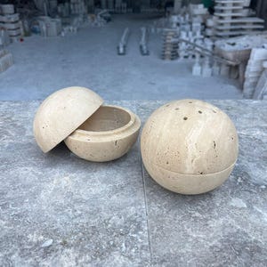 Könnte beinhalten: Zwei beige kugelförmige Behälter aus Stein, einer offen und der andere geschlossen. Der offene Behälter zeigt ein hohles Inneres. Der geschlossene Behälter hat kleine Löcher oben. Die Behälter stehen auf einer grauen Oberfläche, mit einem verschwommenen Hintergrund einer Werkstatt.