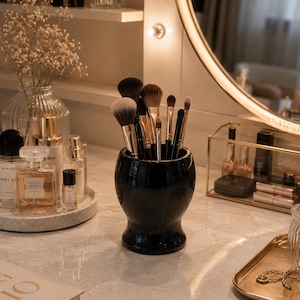 May include: A glossy black ceramic cup filled with various makeup brushes featuring dark and light brown bristles. The cup has a rounded, bulbous shape with a narrow base and sits on a light-coloured marble surface alongside perfume bottles.