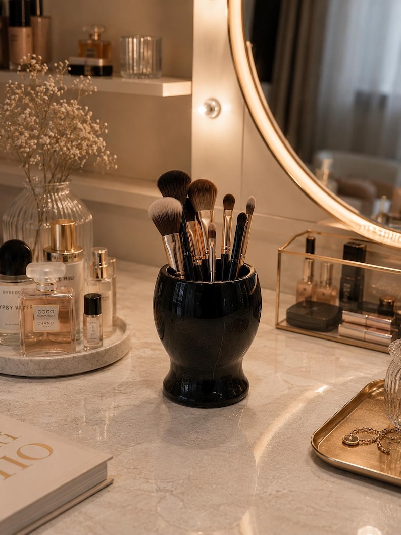 May include: A glossy black ceramic cup filled with various makeup brushes featuring dark and light brown bristles. The cup has a rounded, bulbous shape with a narrow base and sits on a light-coloured marble surface alongside perfume bottles.