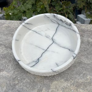 May include: A round, white marble bowl with black veining. The bowl has a textured rim and a smooth interior. This decorative bowl is suitable for serving or display.