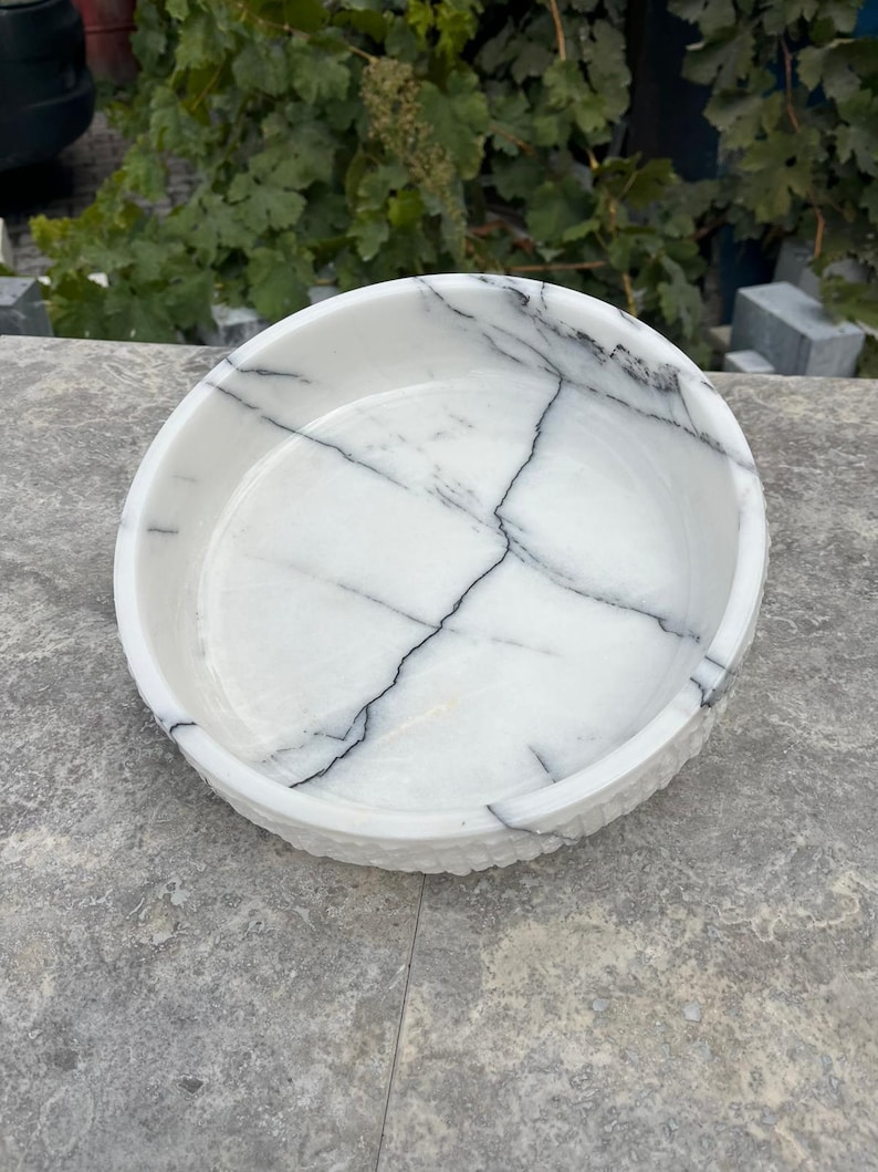 May include: A round, white marble bowl with black veining. The bowl has a textured rim and a smooth interior. This decorative bowl is suitable for serving or display.