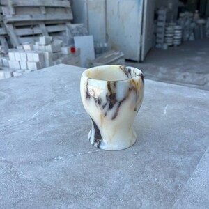 May include: A small, smooth, polished stone cup featuring a creamy off-white base colour with prominent, swirling dark brown and amber veining throughout its surface. The vessel has a rounded, flared rim and a slightly tapered, concave body shape.