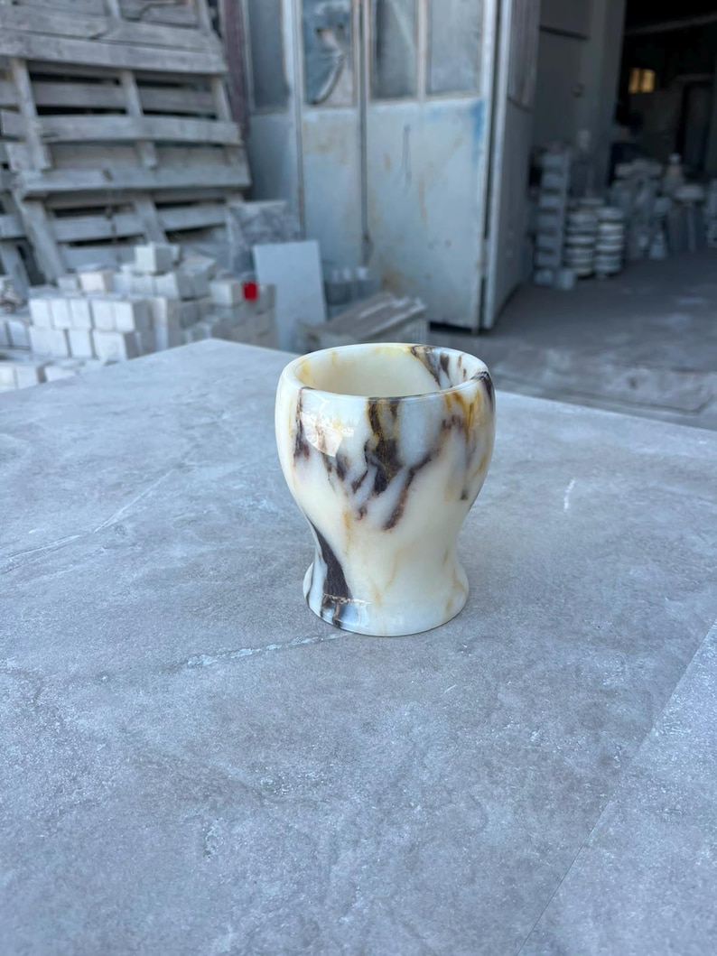 May include: A small, smooth, polished stone cup featuring a creamy off-white base colour with prominent, swirling dark brown and amber veining throughout its surface. The vessel has a rounded, flared rim and a slightly tapered, concave body shape.