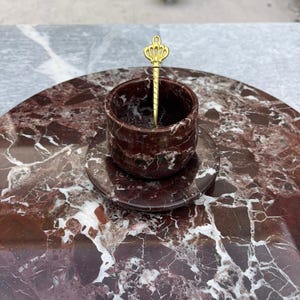 Op de afbeelding: Een decoratieve serveerset met een rond, bordeauxrood marmeren dienblad, een bijpassende kop en schotel. Een goudkleurige lepel met een kroonontwerp rust in de kop. De set is ontworpen voor het serveren van kleine porties.