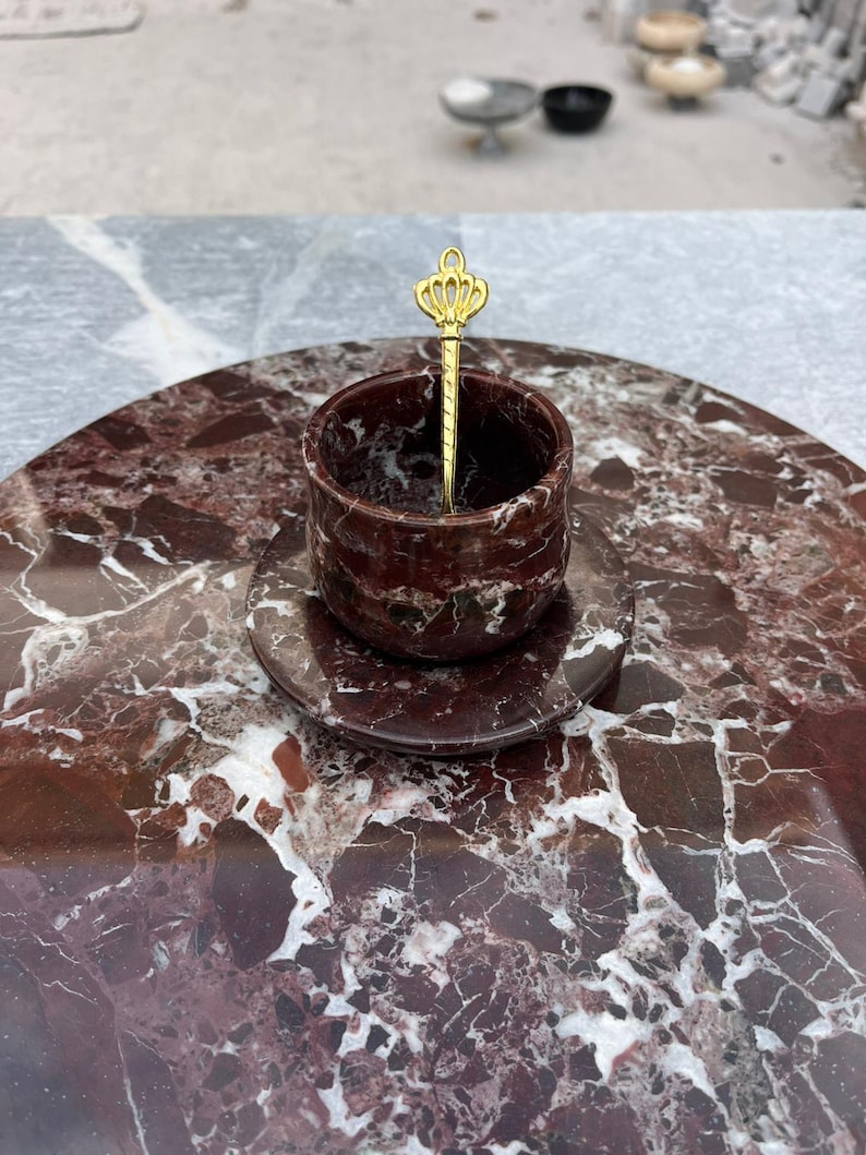 Op de afbeelding: Een decoratieve serveerset met een rond, bordeauxrood marmeren dienblad, een bijpassende kop en schotel. Een goudkleurige lepel met een kroonontwerp rust in de kop. De set is ontworpen voor het serveren van kleine porties.