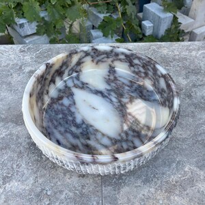 Puede incluir: Un cuenco redondo de m&aacute;rmol con un patr&oacute;n veteado blanco y gris. El cuenco tiene un borde texturizado y estriado. El cuenco est&aacute; sobre una superficie gris.