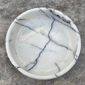 May include: A circular white marble bowl featuring striking black veining patterns. The bowl has a decorative, slightly raised rim. The interior is smooth and polished, highlighting the natural patterns of the stone. A decorative piece.