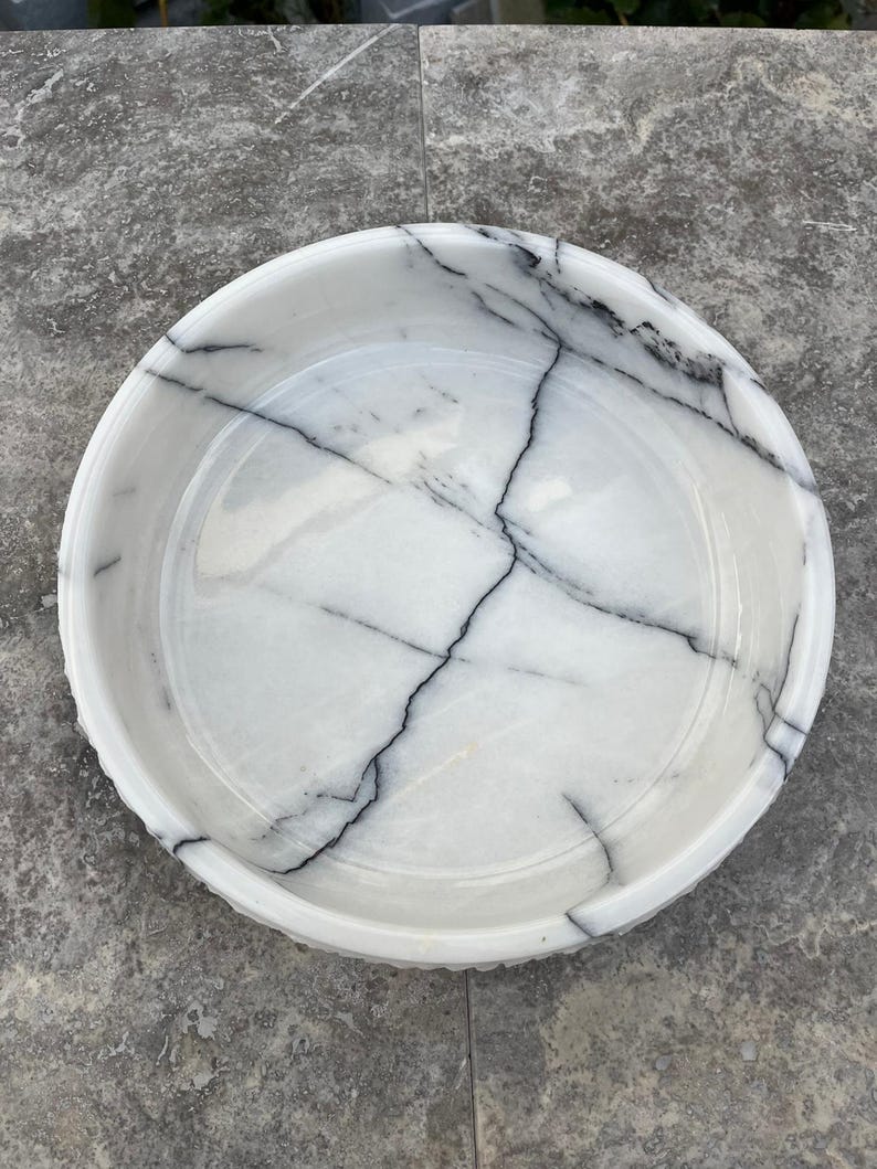 May include: A circular white marble bowl featuring striking black veining patterns. The bowl has a decorative, slightly raised rim. The interior is smooth and polished, highlighting the natural patterns of the stone. A decorative piece.