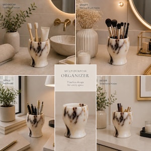 May include: A multipurpose organiser made of white marble with dark brown and grey veining. The cylindrical container features a flared base and a smooth finish, shown holding toothbrushes, makeup brushes, pens, and small decorative items.