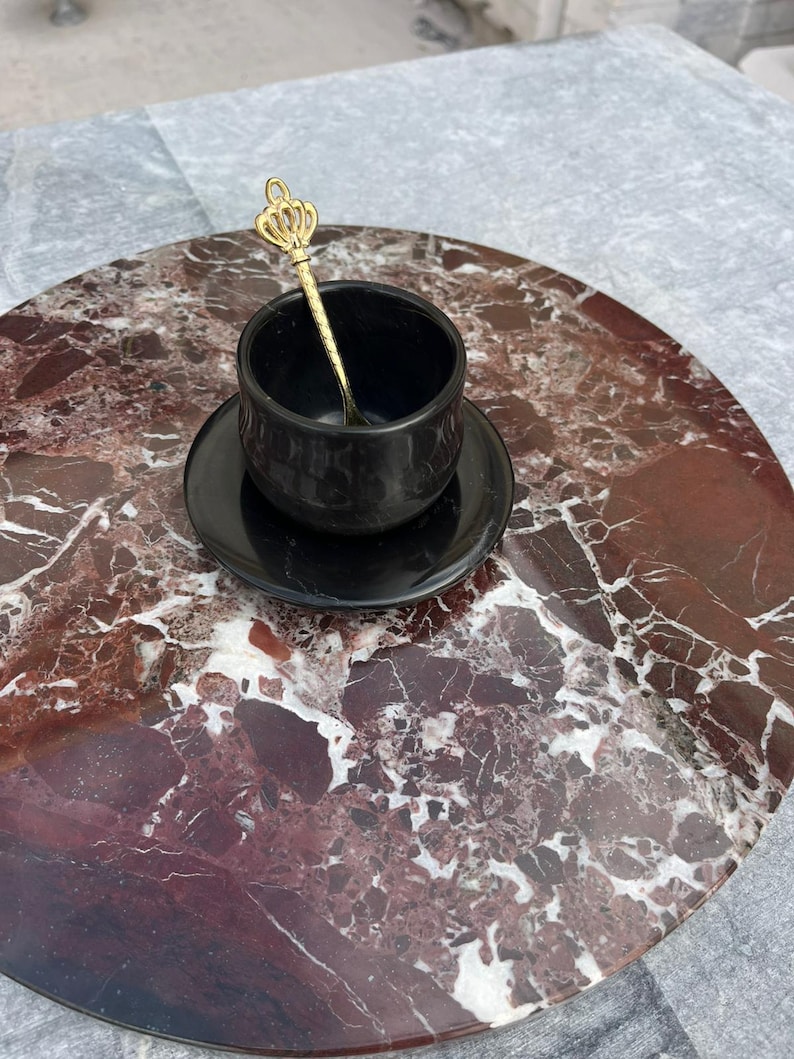 Op de afbeelding: Een zwarte kop en schotel met een gouden kroonvormige lepel, geplaatst op een ronde schaal met een marmerpatroon. De schaal heeft rode, bruine en witte marmering. De achtergrond is een grijs marmeren oppervlak.