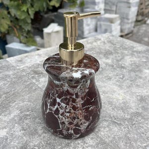 Puede incluir: Un dispensador de jab&oacute;n decorativo con dise&ntilde;o de m&aacute;rmol. El dispensador tiene un patr&oacute;n de m&aacute;rmol rojo oscuro y blanco con una bomba dorada. El dispensador tiene forma redondeada.