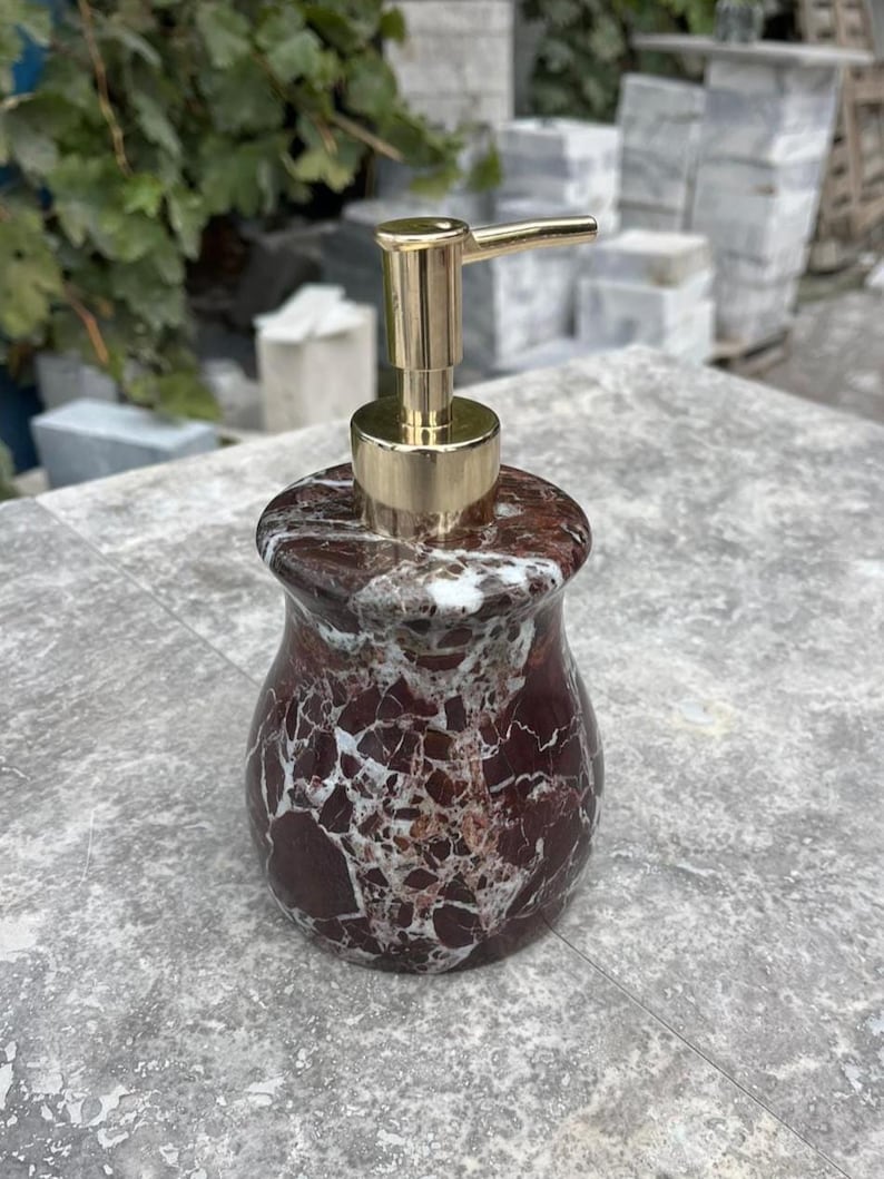Puede incluir: Un dispensador de jab&oacute;n decorativo con dise&ntilde;o de m&aacute;rmol. El dispensador tiene un patr&oacute;n de m&aacute;rmol rojo oscuro y blanco con una bomba dorada. El dispensador tiene forma redondeada.