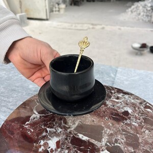 Op de afbeelding: Een zwarte marmeren kop en schotel set met een goudkleurige lepel met een kroonontwerp. De kop staat op een bijpassende schotel en de lepel rust in de kop. De set wordt gepresenteerd op een rood en wit marmeren oppervlak.