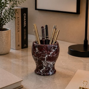 May include: A cylindrical pen holder crafted from deep red marble with prominent white veining, containing a collection of metallic pens. The holder features a flared base and a smooth, polished finish, standing upright on a flat surface.