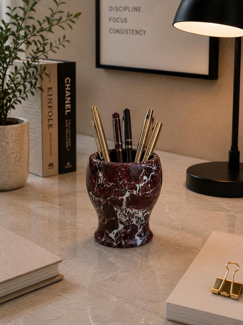 May include: A cylindrical pen holder crafted from deep red marble with prominent white veining, containing a collection of metallic pens. The holder features a flared base and a smooth, polished finish, standing upright on a flat surface.