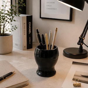 May include: A glossy black ceramic pen holder with a rounded, pedestal-style base, containing five assorted pens in black, gold, and silver tones. The holder is positioned on a desk surface alongside a notebook, books, and a black desk lamp.