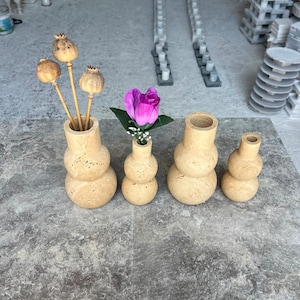 Puede incluir: Cuatro jarrones de piedra travertino beige con una superficie porosa y texturizada y una silueta redondeada apilada. Los dos jarrones m&aacute;s grandes contienen c&aacute;psulas de amapola secas y una rosa artificial morada con hojas verdes.