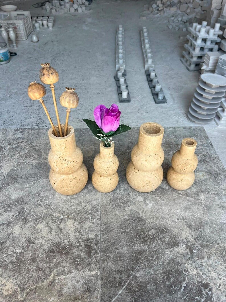 Puede incluir: Cuatro jarrones de piedra travertino beige con una superficie porosa y texturizada y una silueta redondeada apilada. Los dos jarrones m&aacute;s grandes contienen c&aacute;psulas de amapola secas y una rosa artificial morada con hojas verdes.