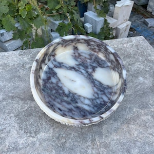 Puede incluir: Un cuenco redondo de m&aacute;rmol con un patr&oacute;n veteado blanco y morado. El cuenco tiene un borde decorativo y acanalado. Est&aacute; sobre una superficie gris. El fondo incluye hojas verdes y bloques de piedra.