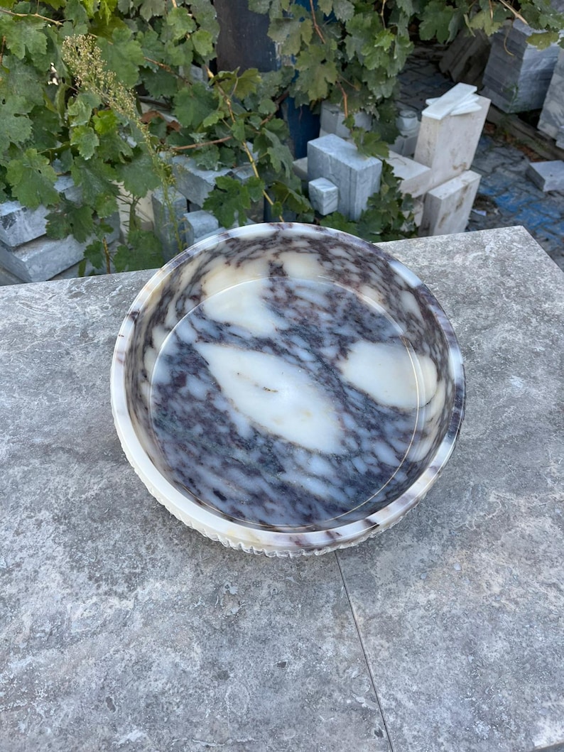 Puede incluir: Un cuenco redondo de m&aacute;rmol con un patr&oacute;n veteado blanco y morado. El cuenco tiene un borde decorativo y acanalado. Est&aacute; sobre una superficie gris. El fondo incluye hojas verdes y bloques de piedra.