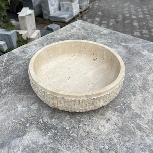 K&ouml;nnte beinhalten: Eine beige, runde Steinschale mit einer strukturierten, geriffelten Au&szlig;enseite. Die Schale besteht aus einem hellfarbigen Stein und steht auf einer grauen Oberfl&auml;che. Das Innere der Schale ist glatt und leer.