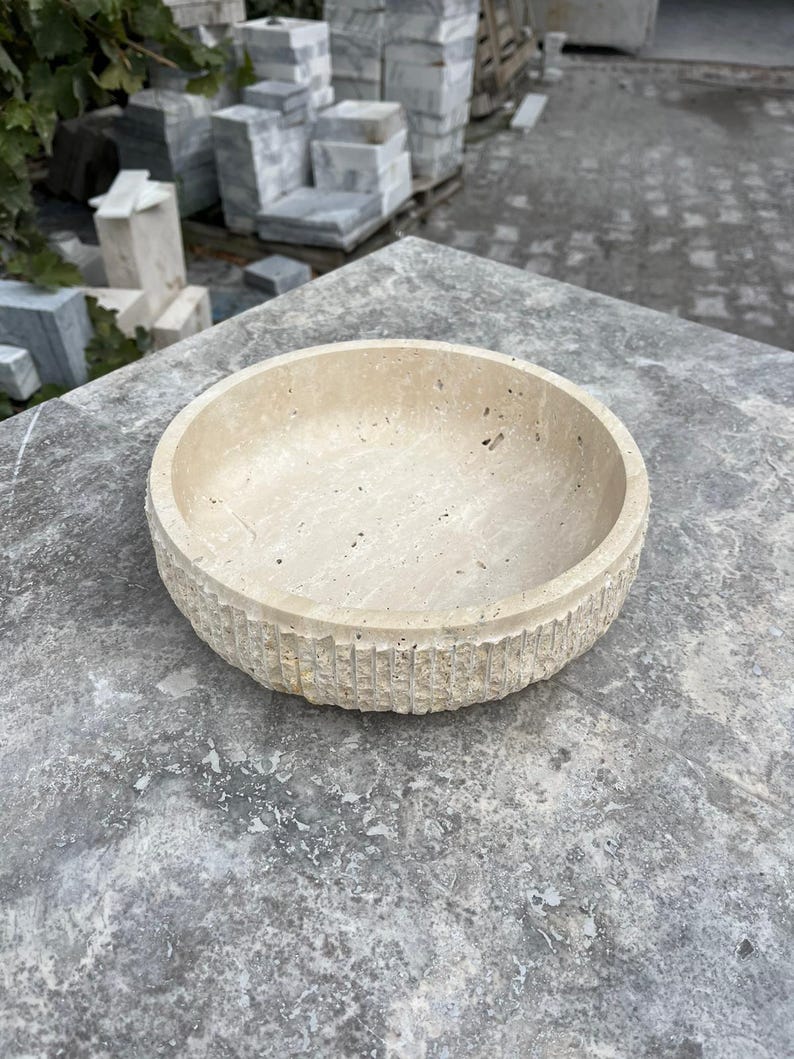 K&ouml;nnte beinhalten: Eine beige, runde Steinschale mit einer strukturierten, geriffelten Au&szlig;enseite. Die Schale besteht aus einem hellfarbigen Stein und steht auf einer grauen Oberfl&auml;che. Das Innere der Schale ist glatt und leer.