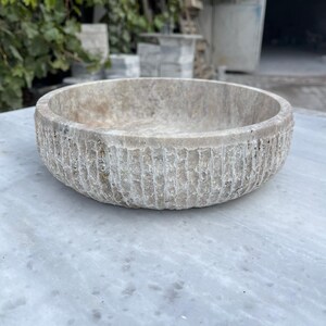May include: A round, stone bowl with a textured, ridged exterior. The bowl is a light beige color and sits on a white marble surface. The bowl's interior is smooth and the overall design is rustic.
