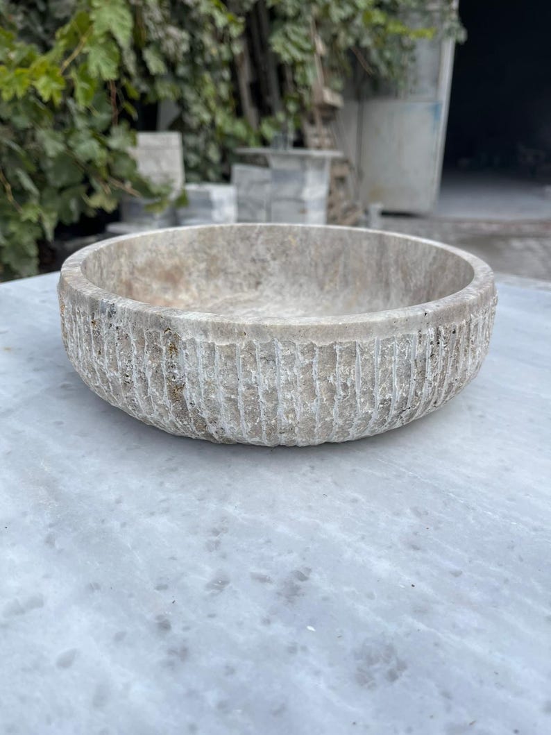 May include: A round, stone bowl with a textured, ridged exterior. The bowl is a light beige color and sits on a white marble surface. The bowl's interior is smooth and the overall design is rustic.