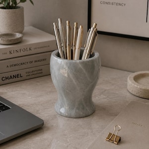 May include: A cylindrical grey marble pen holder with a smooth, polished finish and subtle white veining, containing a collection of various metallic pens with silver and gold-toned clips and barrels, standing upright in a cluster.