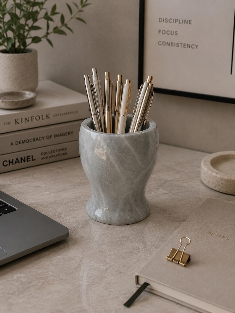 May include: A cylindrical grey marble pen holder with a smooth, polished finish and subtle white veining, containing a collection of various metallic pens with silver and gold-toned clips and barrels, standing upright in a cluster.