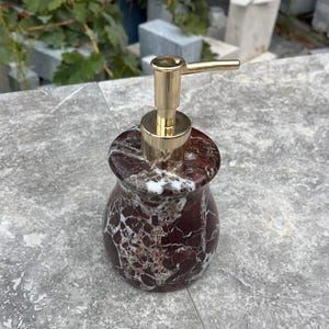 Puede incluir: Un dispensador de jab&oacute;n decorativo con apariencia de m&aacute;rmol. El dispensador tiene un cuerpo con dise&ntilde;o en rojo oscuro y blanco con una bomba dorada. El dispensador est&aacute; dise&ntilde;ado para contener jab&oacute;n l&iacute;quido.