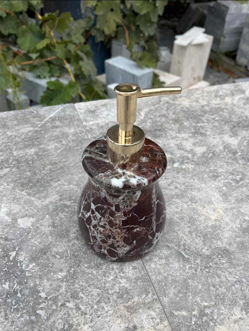 Puede incluir: Un dispensador de jab&oacute;n decorativo con apariencia de m&aacute;rmol. El dispensador tiene un cuerpo con dise&ntilde;o en rojo oscuro y blanco con una bomba dorada. El dispensador est&aacute; dise&ntilde;ado para contener jab&oacute;n l&iacute;quido.